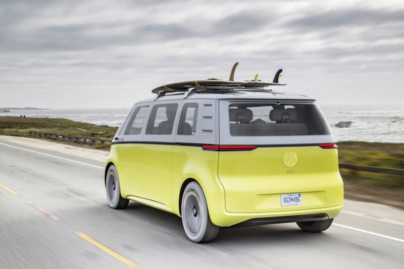 A yellow VW bus concept car drives past the beach, with surfboards on its roof